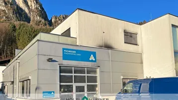 Moderne Gewerbeimmobilie mit grauer Fassade und blauer Beschilderung vor bergiger Landschaft.