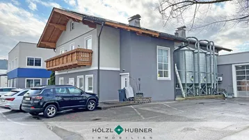 Gebäudeansicht mit traditionellem Balkon und angrenzendem Industriebereich mit Silos und Parkplätzen.
