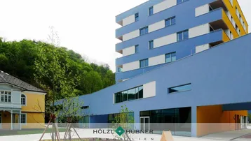 Modernes Gebäude mit blauer Fassade, großen Fenstern und Balkonen, umgeben von Grünflächen.