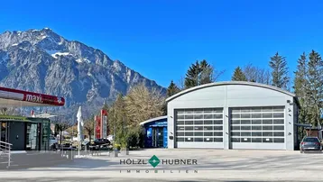 Gewerbegebäude mit zwei großen Garagentoren und einer Tankstelle im Vordergrund, umgeben von Bergen.