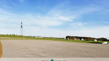 Große, leere Kiesfläche unter blauem Himmel mit Häusern und einem Sendemast in der Ferne.
