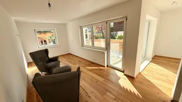 Helles Wohnzimmer mit Holzboden, großen Fenstern und Zugang zu einem Außenbereich.
