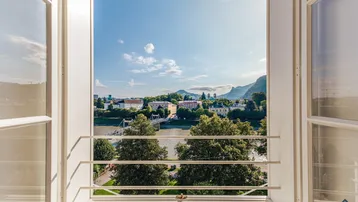 Blick aus einem Fenster auf einen Fluss, eine Stadt und Berge im Hintergrund unter blauem Himmel.