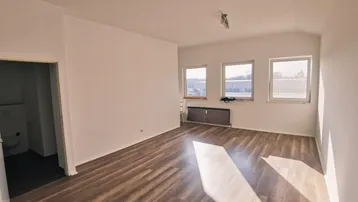 Helles, leeres Zimmer mit Laminatboden und drei Fenstern, das viel Tageslicht hereinlässt.
