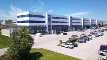 Modernes Bürogebäude mit weißer Fassade und blauen Akzenten, umgeben von einem großen Parkplatz und Grünflächen unter blauem Himmel.