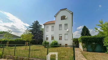 Weißes Einfamilienhaus mit großem Garten und grüner Hecke unter blauem Himmel.