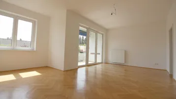 Helles, leeres Wohnzimmer mit Fischgrätparkett und Zugang zu einem Balkon.