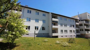 Mehrfamilienhaus mit weißer Fassade und Grünfläche im Vordergrund unter blauem Himmel.