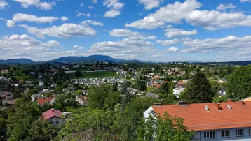 Weitläufiger Blick über eine grüne Wohnlandschaft mit Bergen im Hintergrund unter blauem Himmel.