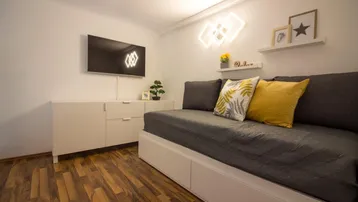Helles Wohnzimmer mit modernem Tagesbett, TV und Holzfußboden, akzentuiert durch Neonbeleuchtung.