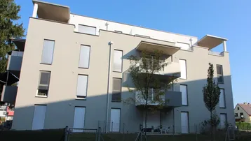 Modernes Mehrfamilienhaus mit heller Fassade, Balkonen und großen Fenstern unter blauem Himmel.