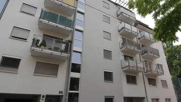 Mehrfamilienhaus mit mehreren Balkonen und Rollläden an den Fenstern, umgeben von Grün.
