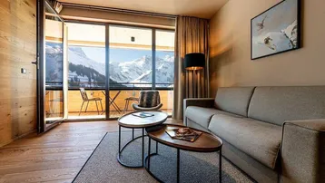 Gemütliches Wohnzimmer mit Holzboden, großem Fenster und atemberaubendem Bergblick von einem Balkon.