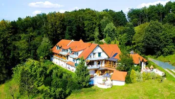 Großes Anwesen mit mehreren Gebäuden, umgeben von Wald und grünen Hügeln, mit roten Ziegeldächern.