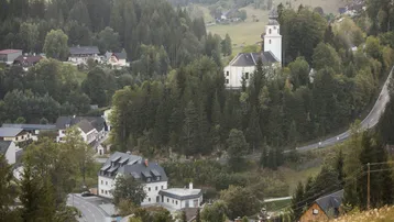 Malerische Landschaft mit Häusern, Wald und einer Kirche, eingebettet in eine hügelige Umgebung.