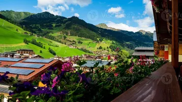 Blick von einem Balkon mit Blumenkästen auf ein Bergdorf und grüne Hänge mit Solaranlagen auf den Dächern.