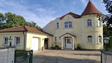 Gelbe Villa mit Turm, gepflasterter Auffahrt und integrierter Garage unter blauem Himmel.