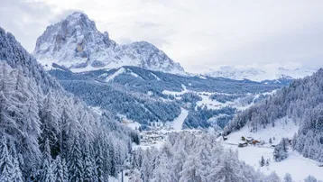 Winterliche Berglandschaft mit schneebedeckten Bäumen, einem Tal und einem kleinen Dorf, umgeben von majestätischen Gipfeln.