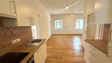 Blick von der Küche in einen hellen Wohnbereich mit Holzboden und kleinen Fenstern.