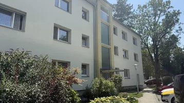 Mehrfamilienhaus mit heller Fassade und gepflegter Grünanlage unter blauem Himmel.