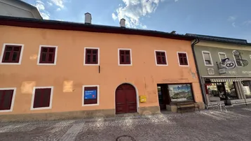 Vorderansicht eines historischen Gebäudes mit orangefarbener Fassade und roten Fensterläden an einer gepflasterten Straße.
