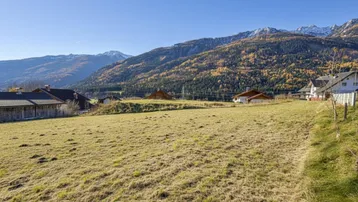 Weitläufige Wiesenfläche mit Blick auf die umliegende Berglandschaft und vereinzelte Häuser unter blauem Himmel.