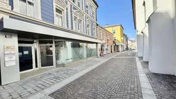 Blick auf die gepflasterte Straße und die historische Fassade des Gebäudes mit Geschäftsräumen im Erdgeschoss.