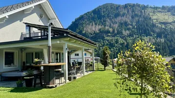 Haus mit überdachter Terrasse und großem Garten vor beeindruckender Bergkulisse.