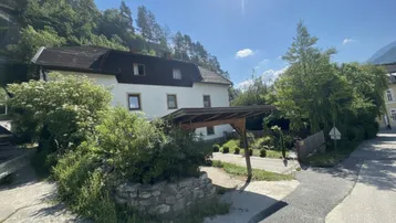 Hausfassade mit Carport und üppiger Vegetation vor einer malerischen Bergkulisse an einem sonnigen Tag.