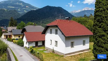 Weißes Haus mit rotem Ziegeldach in einer ländlichen Umgebung mit Bergen im Hintergrund.
