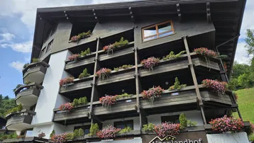Alpenländisches Gebäude mit vielen Balkonen, geschmückt mit bunten Blumenkästen vor grüner Bergkulisse.