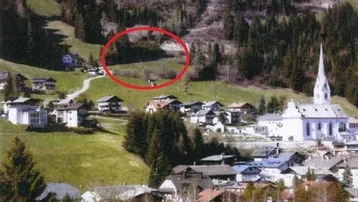 Luftaufnahme eines Dorfes in den Bergen, mit einem rot markierten Baugrundstück am Sonnenhang.