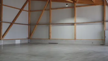 Große, leere Halle mit Betonboden, weißen Wellblechwänden und sichtbarer Holzkonstruktion im Dachbereich.