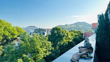 Weitblick von einem Balkon über grüne Bäume und Dächer auf die umliegende Hügellandschaft.