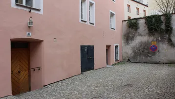 Ansicht eines rosafarbenen Gebäudes mit Kopfsteinpflasterhof und einer alten Holztür, umgeben von einer Mauer.