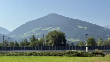 Panoramablick auf eine bewaldete Berglandschaft mit einem grünen Feld im Vordergrund und einer Lärmschutzwand.