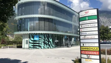 Modernes Geschäftsgebäude mit geschwungener Glasfassade und einem großen Hinweisschild vor einem Bergpanorama.