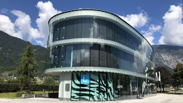 Modernes Gebäude mit geschwungener Glasfassade und großen Fensterflächen, eingebettet in eine Berglandschaft.
