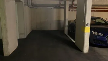 Tiefgaragenstellplatz mit Betonwänden und sichtbaren Leitungen, ein blauer Sportwagen ist teilweise geparkt.