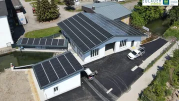 Luftaufnahme eines modernen Gebäudes mit Photovoltaikanlage auf dem Dach und gepflastertem Vorplatz am Wasser.