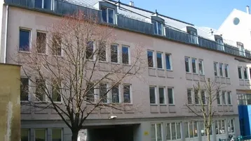 Mehrstöckiges Gebäude mit zahlreichen Fenstern, Dachgauben und einer überdachten Einfahrt im Erdgeschoss.
