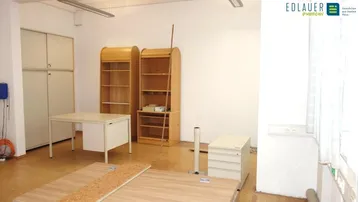 Geräumiges Büro mit mehreren Schreibtischen, Holzregalen und Aktenschränken, gut beleuchtet durch Deckenleuchten und Fenster.