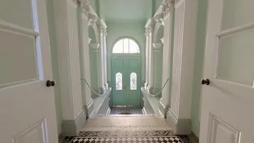 Großzügiger Eingangsbereich mit hohen Decken, Säulen und einem Schachbrettmuster-Fliesenboden, der zum Haupteingang führt.