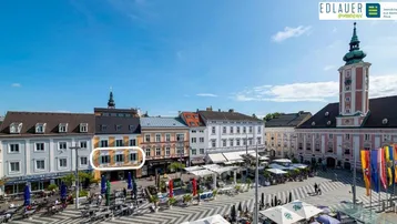 Luftaufnahme eines belebten Stadtplatzes mit historischen Gebäuden und einem markanten Kirchturm.