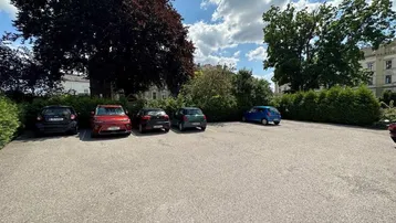 Geräumiger Außenparkplatz mit mehreren Fahrzeugen unter schattenspendenden Bäumen an einem sonnigen Tag.
