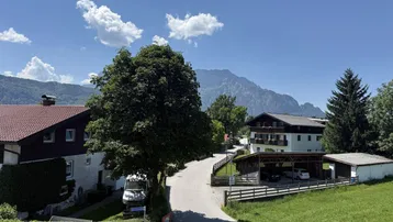 Blick auf eine ländliche Straße mit Häusern, umgeben von grünen Hügeln und Bergen unter blauem Himmel.