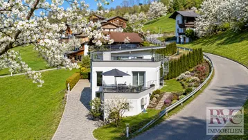Modernes Haus mit mehreren Terrassen und gepflegter Gartenanlage in einer grünen Hügellandschaft.