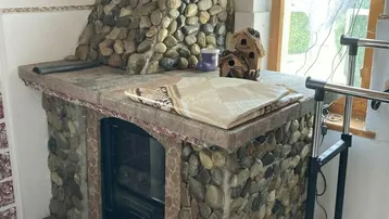 Rustikaler Kamin aus Flusssteinen mit einem Holzofen und einem Fenster mit Vorhängen.