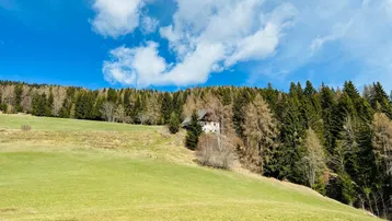 Haus in einer weiten, grünen Wiesenlandschaft, umgeben von Wald und Bergen unter blauem Himmel.