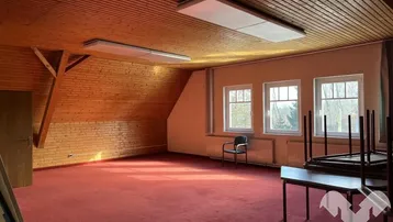 Großer Mehrzweckraum im Dachgeschoss mit Holzwänden, Holzdecke und rotem Teppichboden, hell durch mehrere Fenster.